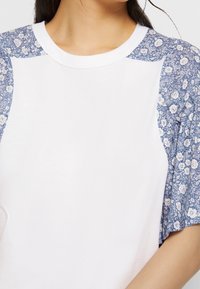 Camiseta blanca de mangas cortas con mangas raglán de estampado floral azul y cuello redondo. La tela parece suave con una textura lisa.