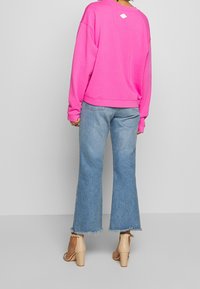 Sudadera rosa de corte relajado con puños ribeteados, combinada con jeans de mezclilla claros de pierna acampanada con dobladillos deshilachados. La modelo lleva sandalias de tacón alto color beige.
