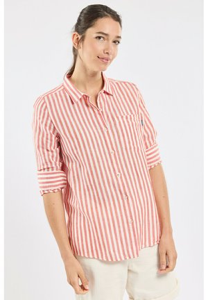 Femme portant une chemise à rayures verticales roses et blanches à boutons, manches retroussées, et un pantalon de couleur claire, debout devant un fond uni.