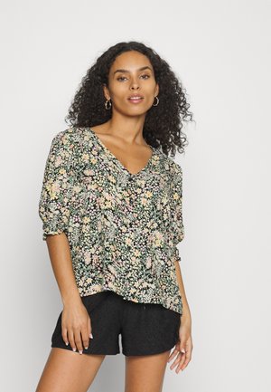 Blusa floral com decote em V, apresentando mangas curtas bufantes, fundo preto com padrões de flores coloridas. Usada com calções pretos de rendinha.