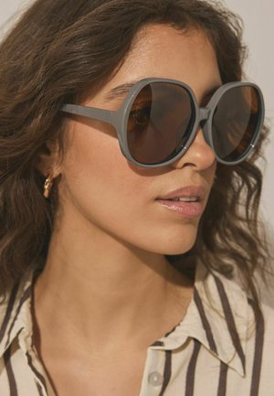 Kvinde med mellemtonet hud, der bærer store gråstel solbriller, guldfarvet ørering og en cremefarvet skjorte med mørke lodrette striber.