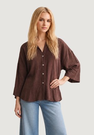 Vrouw met lang blond haar draagt een losse bruine blouse met knopen en lichtblauwe jeans, staand met één hand in haar zij tegen een effen achtergrond.
