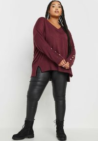 Bordeauxfarbener übergroßer Pullover mit V-Ausschnitt, gerippter Textur und seitlichen Schlitzen, mit metallischen Knopfakzenten an den Ärmeln, kombiniert mit schwarzen, eng anliegenden Lederhosen.