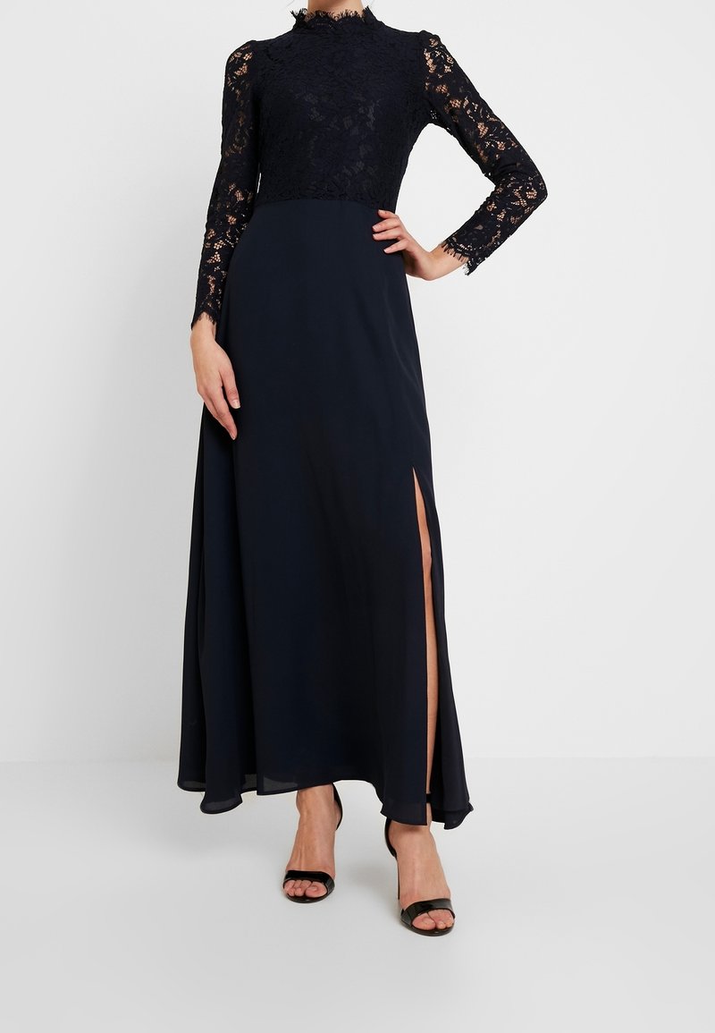 Femme portant une longue robe noire avec des manches longues en dentelle et une fente haute, posant avec une main sur la hanche et des talons ouverts noirs.