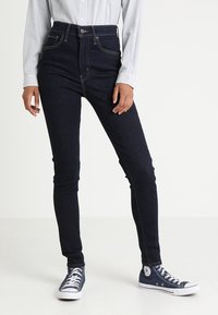 Vaqueros de mezclilla oscuros de corte slim con cintura alta, costuras en contraste, diseño de cinco bolsillos y cierre de botón, combinados con zapatillas de lona azules.