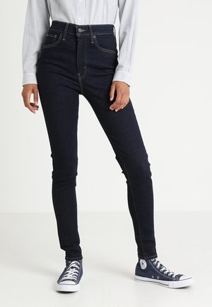Vaqueros de mezclilla oscuros de corte slim con cintura alta, costuras en contraste, diseño de cinco bolsillos y cierre de botón, combinados con zapatillas de lona azules.