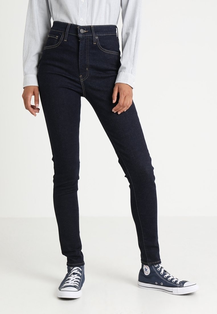 Vaqueros de mezclilla oscuros de corte slim con cintura alta, costuras en contraste, diseño de cinco bolsillos y cierre de botón, combinados con zapatillas de lona azules.