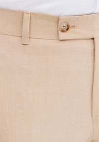 Beżowe spodnie dopasowane o gładkiej teksturze, z zapięciem na guzik i szlufkami na pasek. Materiał wydaje się być wytrzymały i lekko teksturowany.