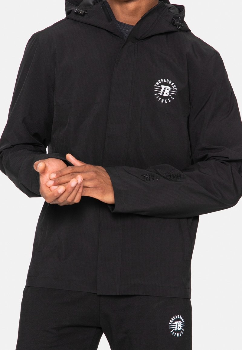 Schwarze leichte Jacke mit Kapuze, strukturiertem Stoff, Reißverschluss vorne und weißem Logo auf der linken Brustseite. Verfügt über elastische Bündchen und versteckte Taschen.