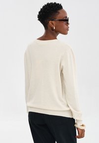 Sweter w kolorze kremowym, z długim rękawem, o miękkiej teksturze, luźnym kroju, z ribbowanym wykończeniem na mankietach, zestawiony z czarnymi spodniami, widok z tyłu.