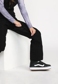Pantalons de sport noirs avec poches zippées, associés à des baskets noires avec des accents blancs et des semelles en caoutchouc gomme. Haut à manches longues violet clair.