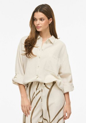 Kvinne med langt hår iført en løs beige skjorte med knapper og beige bukser med brune abstrakte mønstre, stående mot en hvit bakgrunn.