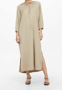 Lange beige jurk gemaakt van lichtgewicht stof, met een geraffineerde halslijnen, strikdetail en zijsplitten. Lange mouwen eindigen bij de pols.