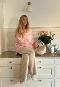 Sieviete rozā trikotētā džemperī sēž uz virtuves letes, maisot krēmkrāsas bļodā, apkārt viņai ir rozeņu pušķis.
