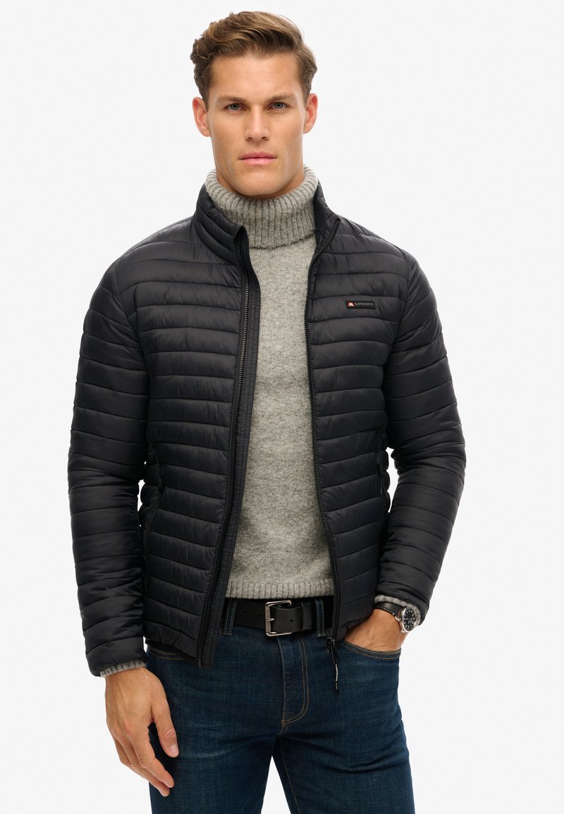 Chaqueta negra acolchada con cuello alzado, combinada con un suéter de cuello alto gris claro. Cuenta con un cierre de cremallera y texturas contrastantes.