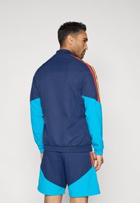 Ropa deportiva para hombres que incluye una chaqueta deportiva azul marino con mangas turquesas y detalles rojos, combinada con pantalones cortos a juego. Textura ligera y suave.