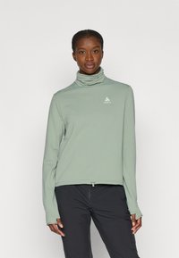 Lys grøn langærmet top med en mock neck, struktureret stof og et lille logo. Har en indsnævret kant og tommelhuller i ærmerne.