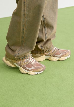 Zapatillas en malla beige y marrón con detalles en blanco; parte superior texturizada, suela acolchada y detalle de logotipo prominente; combinadas con unos vaqueros verdes holgados.