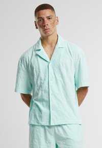Camisa de manga corta azul claro en tejido acolchado con un patrón en relieve. Cuenta con un cuello, cierre de botones en el frente y un discreto detalle de logo.