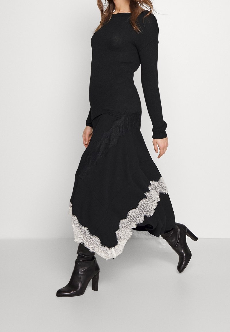 Femme portant un haut noir à manches longues, une jupe noire avec un bord en dentelle blanche, et des bottines noires à talons, marchant devant un fond blanc uni.
