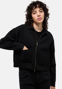 Veste noire courte à fermeture éclair en tissu doux, avec un col, deux poches avant et une finition texturée. La fermeture éclair est équipée d'un tire-zip en métal.