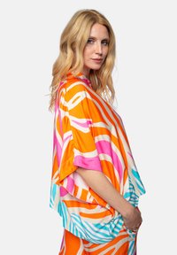 Kolorowa bluzka w stylu kimono w intensywnych wzorach pomarańczowym, różowym i turkusowym. Miękka tkanina, luźny krój, szerokie rękawy i abstrakcyjny wzór.