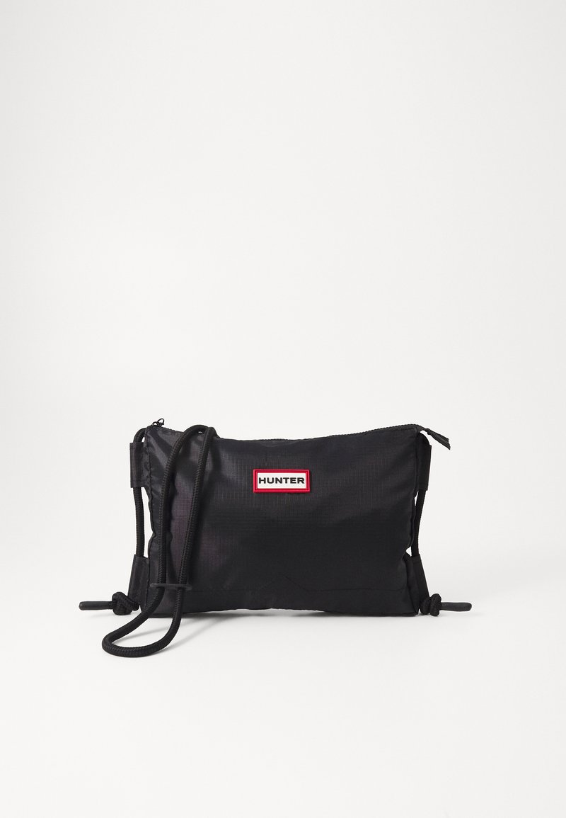 Bolsa tiracolo preta em nylon com formato retangular, alças ajustáveis com cordão e um emblema em vermelho e branco. Fecho com zíper.