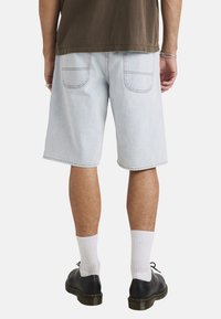 Pantalones cortos de mezclilla de color azul claro con un ajuste holgado, que cuentan con dos bolsillos traseros y costuras en contraste. Combinados con zapatos negros y calcetines blancos.