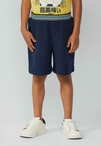 Pantaloni corti blu navy con cintura elastica e dettagli a righe. Indossati con sneakers bianche e una t-shirt grafica gialla.