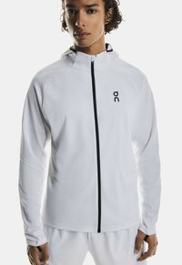 White zip-up athletic jacket with a hood, featuring black zipper and logo. Smooth texture, long sleeves, and a fitted design.