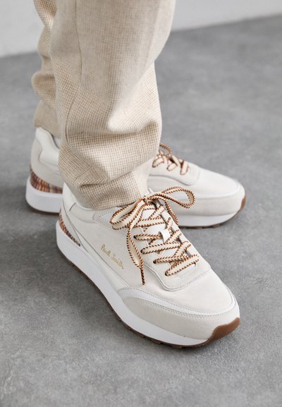 Tenisi albi din suede cu accente cu model, șireturi maro și o talpă din cauciuc texturată. Scrisă de marcă aurie pe lateral.