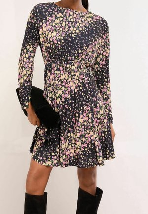 Mujer que lleva un vestido largo de manga larga negro con flores rosas y amarillas, sosteniendo un bolso de mano negro y llevando botas negras hasta la rodilla.