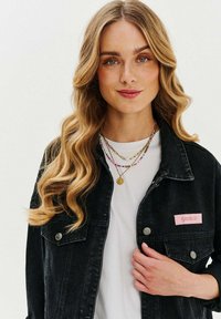 Czarna kurtka dżinsowa z różową naszywką z napisem "GIRLY". Posiada srebrne akcesoria, klasyczny kołnierz i zapięcia na guziki. Nosi ją na białej koszuli.