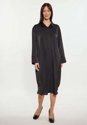 Frau steht, trägt ein schwarzes Satin-Midikleid mit langen Ärmeln, Knöpfen, Kragen und schwarzen Absätzen vor weißem Hintergrund.