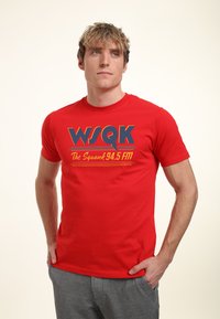 Joven con camiseta roja de WSQK The Squawk 94.5 FM, de pie con las manos en los bolsillos, mirando ligeramente hacia arriba y a un lado.