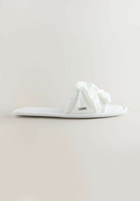 Valge pehme polsterdatud suss pehme, voltidega ülaosaga ja väike metallist brändisilt tavalise heleda tausta peal.