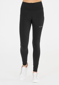 Mustat legginsit korkealla vyötäröllä, istuva siluetti ja sileä pinta; sisältävät pienen valkoisen logon alhaalla. Yhdistetty valkoisiin tennareihin.