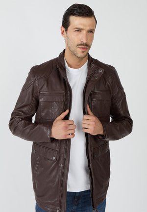 Braune Lederjacke mit Reißverschluss vorne, hohem Kragen und vier vorderen Taschen. Glatte Textur mit dezenten Nähten. Über einem weißen T-Shirt getragen.