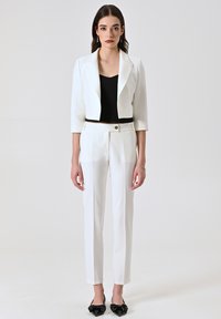 Blazer blanc court sur un haut noir, associé à un pantalon blanc sur mesure. Des ballerines pointues noires complètent la tenue. Tissu lisse, design structuré.