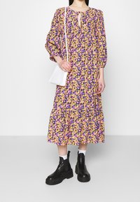 Blommig maxikjol med lila, rosa och gula mönster, nyckelhålsringning och puffärmar. Ovanpå bärs svarta platåstövlar och en vit väska.