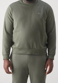 Bluza w oliwkowej zieleni wykonana z miękkiej bawełnianej mieszanki, z okrągłym dekoltem, długimi rękawami, ściągaczami na mankietach oraz małym haftowanym logo na piersi.