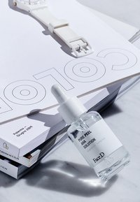 Una bottiglia contagocce di vetro trasparente etichettata "Pure Peel AHA Solution" si trova su un mucchio di riviste insieme a un orologio bianco e texture morbide.