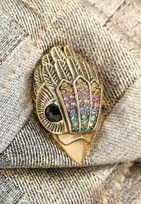 Broche en forme d'oiseau en ton or, ornée d'accents de gemmes colorées sur le corps et d'un œil en pierre noire. Fond en tissu texturé.