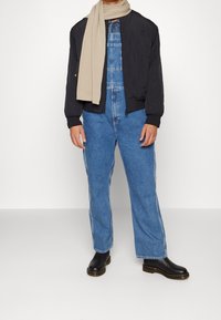 Černá bomber bunda přes denní overaly, světle béžový šátek přehozený, volné modré džíny a černé boty se silnou podrážkou.