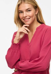 Femme blonde souriante, main près du menton, portant des boucles d'oreilles créoles dorées et un chemisier rose framboise texturé à manches longues avec des poignets froncés.