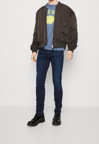 Chaqueta bomber marrón, camiseta gráfica azul con diseño amarillo, jeans oscuros ajustados de mezclilla y botas de cuero negras con cordones.
