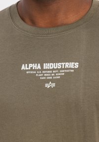 Prim-plan al unei cămăși de culoare verde-oliv cu text alb: "ALPHA INDUSTRIES," detalii ale contractorului Departamentului de Apărare al SUA și un logo stilizat "A".