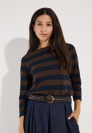 Vrouw met lang donker haar die een bruin en marineblauw gestreept topje, donkere geplooide broek en een geweven riem met een ronde gesp draagt.