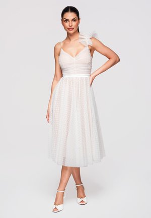 Femme portant une robe midi blanche avec une bretelle à volants transparente et un tissu texturé à pois, associée à des talons blancs.