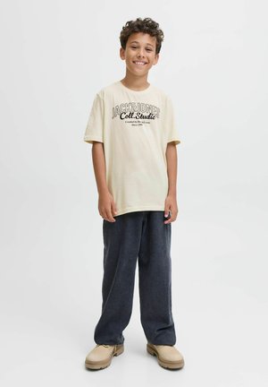 Helles Gelbes T-Shirt mit schwarzem Textlogo auf der Vorderseite, kombiniert mit locker sitzenden dunklen Denim-Hosen und beigen Schuhen.